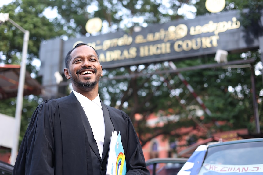 A young Indian man in barrister robes standing outside a court, smiling 