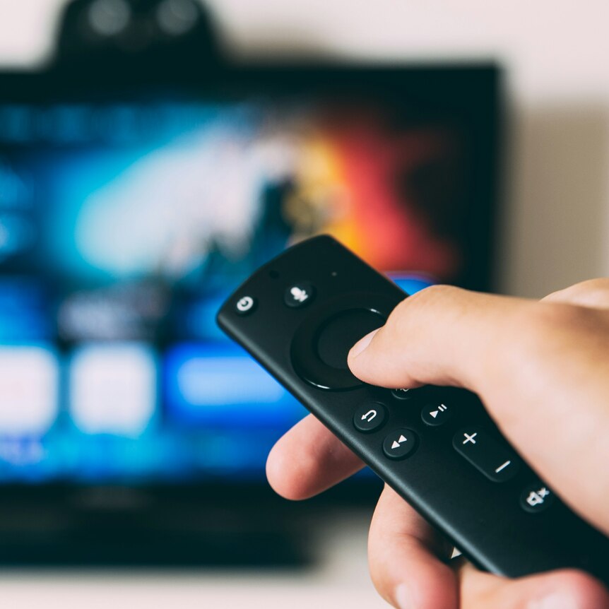 A close-up of a hand holding a small television remote, which is pointed at an out-of-focus TV.