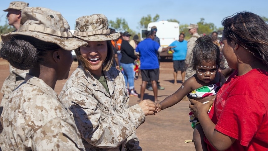 Two smiling women in US military uniform shake the hand of an Indigenous toddler wearing cultural face paint