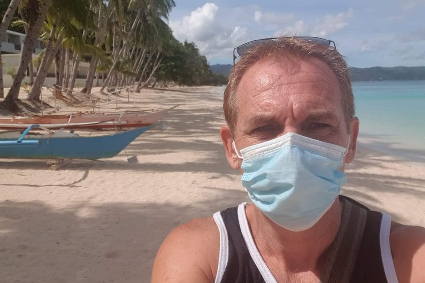 A man looks into the camera as he wears a face mask. A beach is seen behind him with boats to the left.