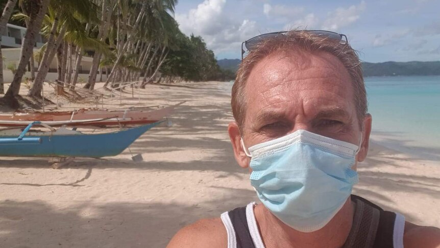 A man looks into the camera as he wears a face mask. A beach is seen behind him with boats to the left.