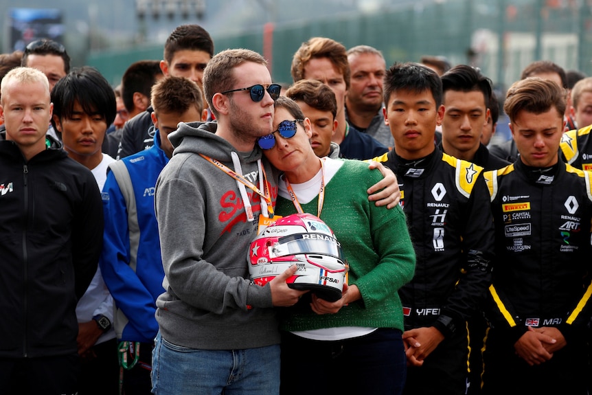 Anthoine Hubert's brother and mother hold the late driver's helmet and each other in front of sombre looking drivers