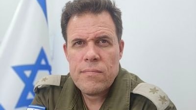 A man staring intensely at the camera wearing army gear with an Israeli flag in the background