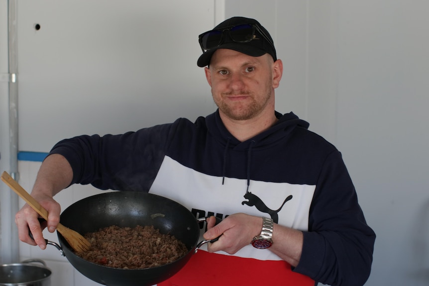 Man in cap looking at camera holding pan full of cooked mince.
