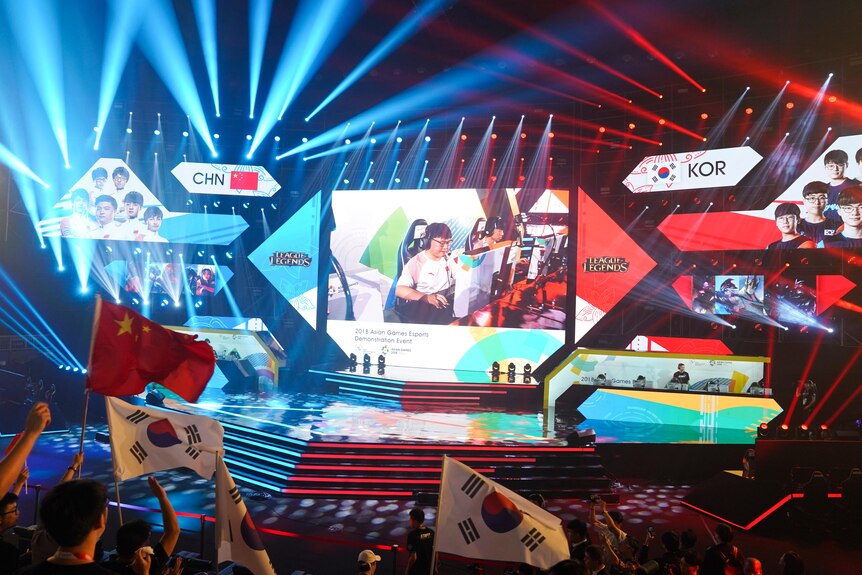Players in an esports competition are shown on a big screen in an auditorium as fans wave Chinese and South Korean flags.