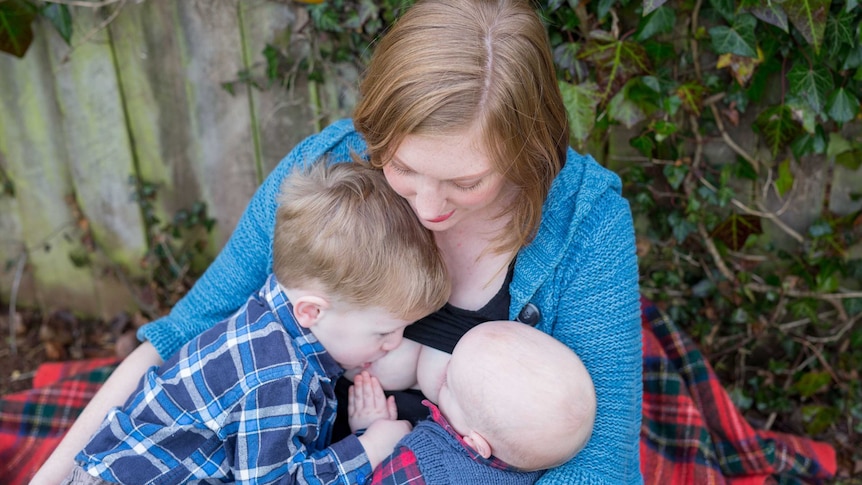 A woman with a blue cardigan breastfeeding a two babies at the same time with no faces fully visible.