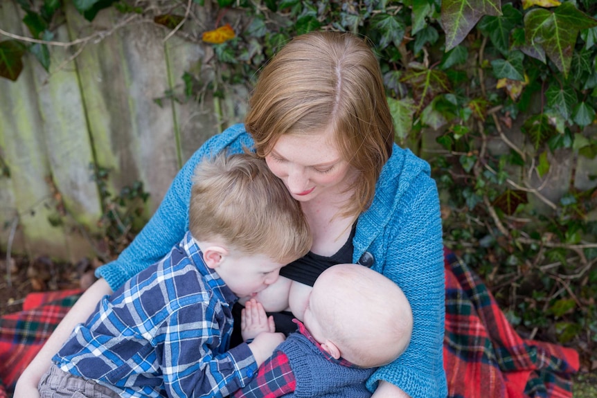 A woman with a blue cardigan breastfeeding a two babies at the same time with no faces fully visible.