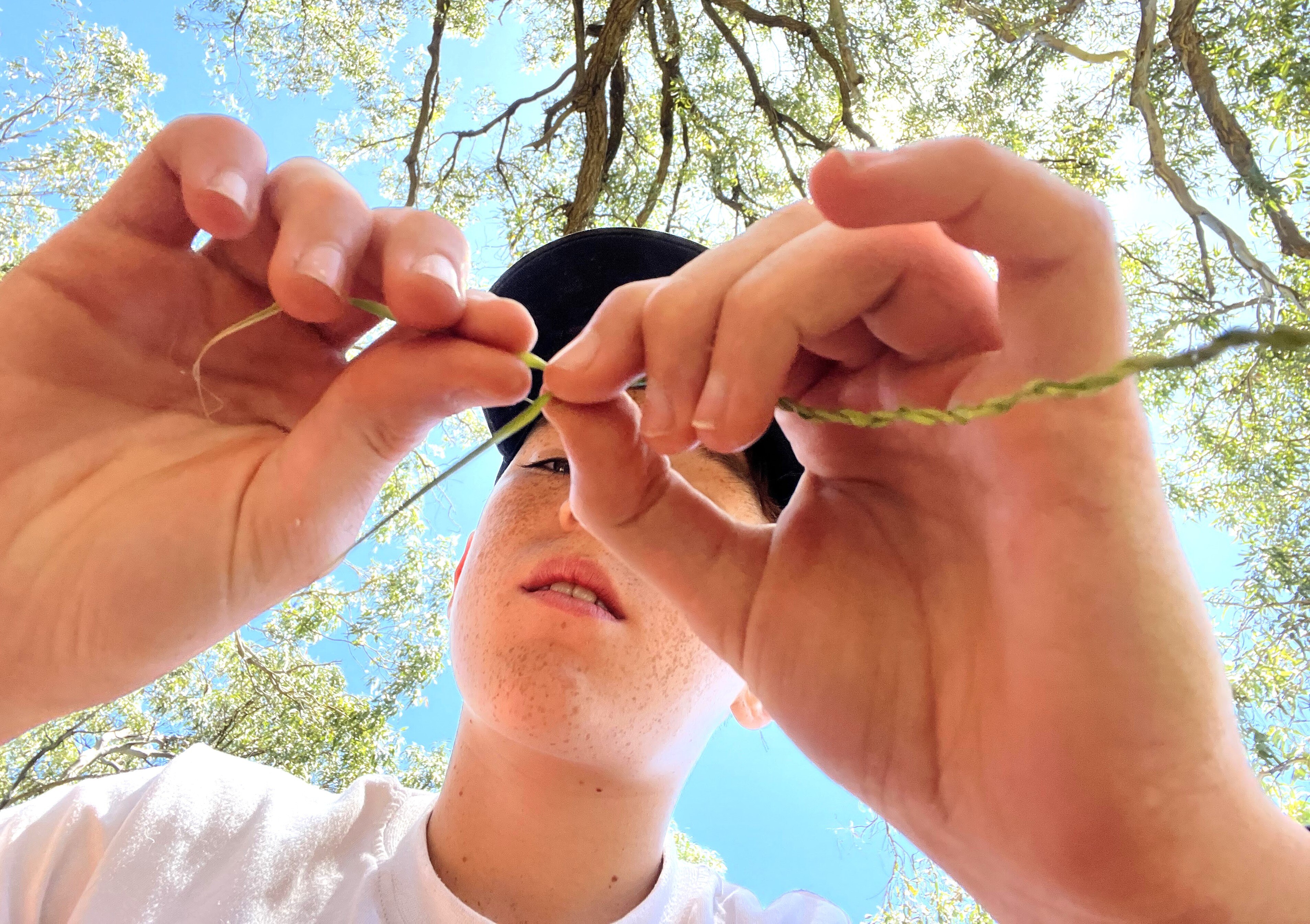 A young boy wearing a white shirt and black cap closely ties a knot out of leafy material under a tree.