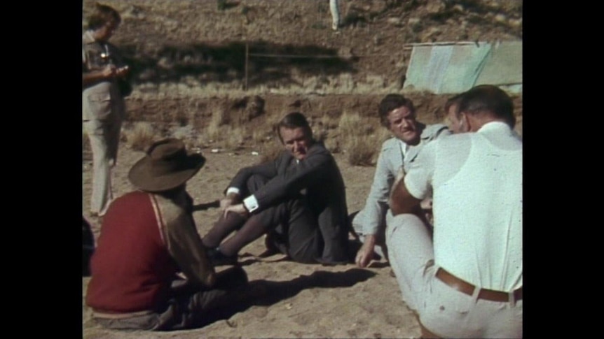 four men sitting on the sand having a discussion, one of which is Malcolm Fraser
