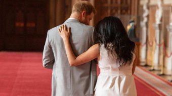 Prince Harry, in a grey suit, holds baby Archie as Meghan, in white, has her arm around him. The pair are walking away.