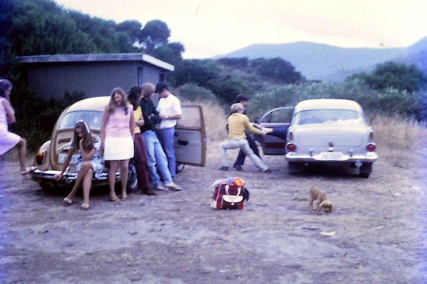Colour photo from the 1970s of 6 people leaning on a honey brown VW beetle, and 2 boys pulling a person out of the seat of car.