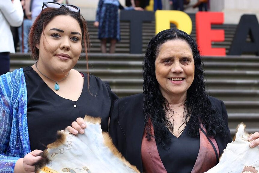 Nakia Cadd and Muriel Bamblett smile as they hold a possum-skin cloak together on the steps of Parliament House.
