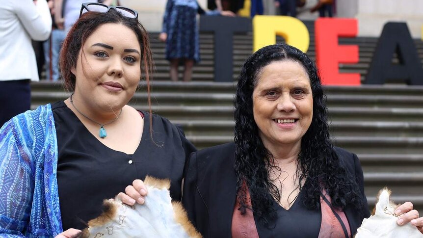 Nakia Cadd and Muriel Bamblett smile as they hold a possum-skin cloak together on the steps of Parliament House.
