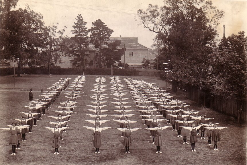 Women stand in lines with their arms out, doing exercises on the lawn of a school in the 1920s.