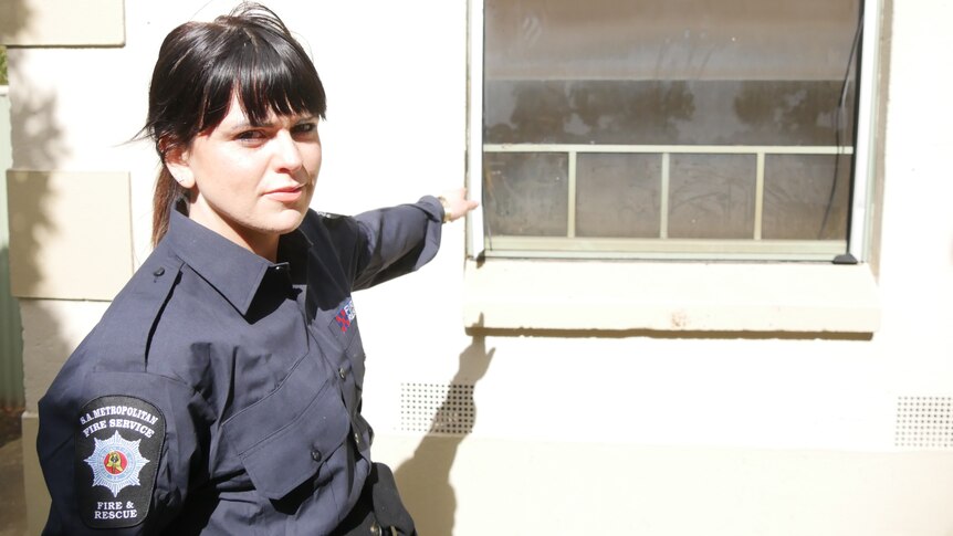 A woman in an emergency services uniform stands next to an outside wall pointing at a window