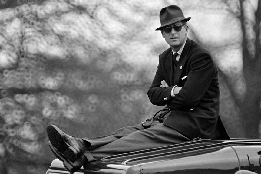 Black and white photo of Prince Philip sitting atop a car, wearing a suit, hat and sunglasses, in 1965.