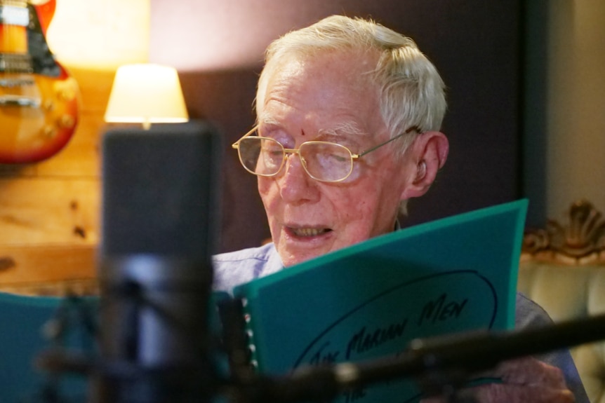 an older man with glasses on looks down at a blue folder, with a microphone in front of his face