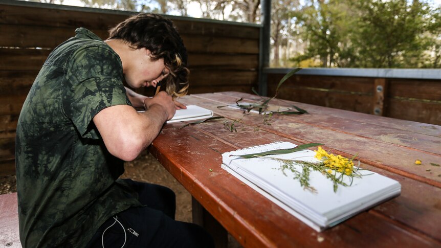 Bailey Muscat sitting at a picnic table in the bush drawing Indigenous flora.