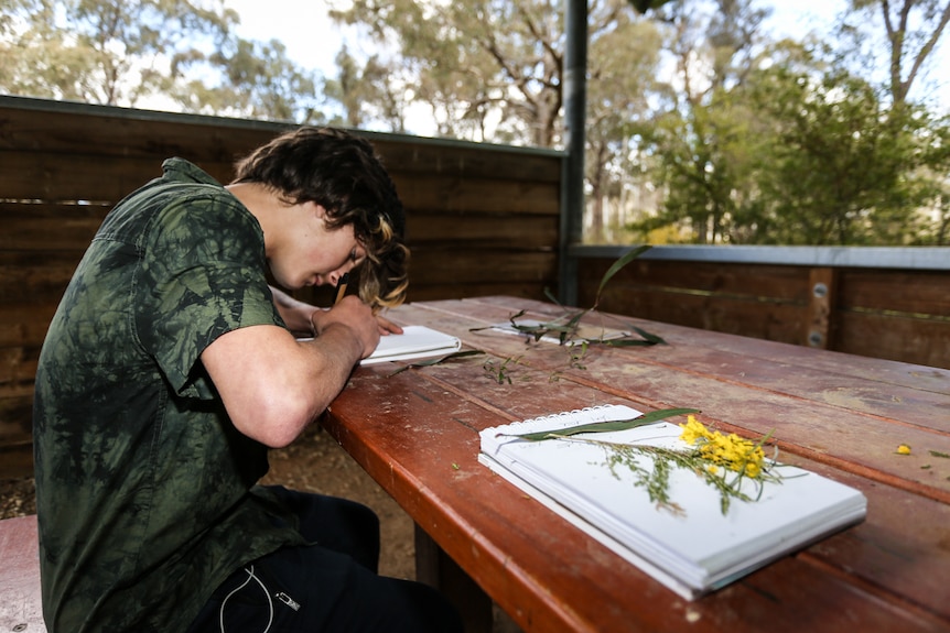 Bailey Muscat sitting at a picnic table in the bush drawing Indigenous flora.