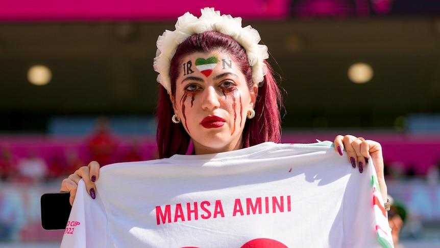 An Iran fan wears makeup showing tears while holding a white shirt with MAHSA AMINI written on it