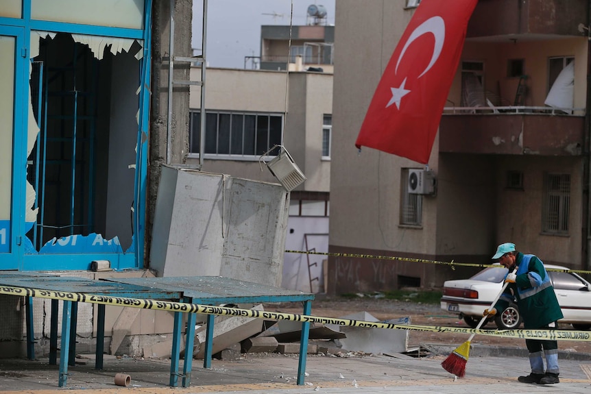 Wide shot of a man sweeping debris on the ground next to a damaged building.