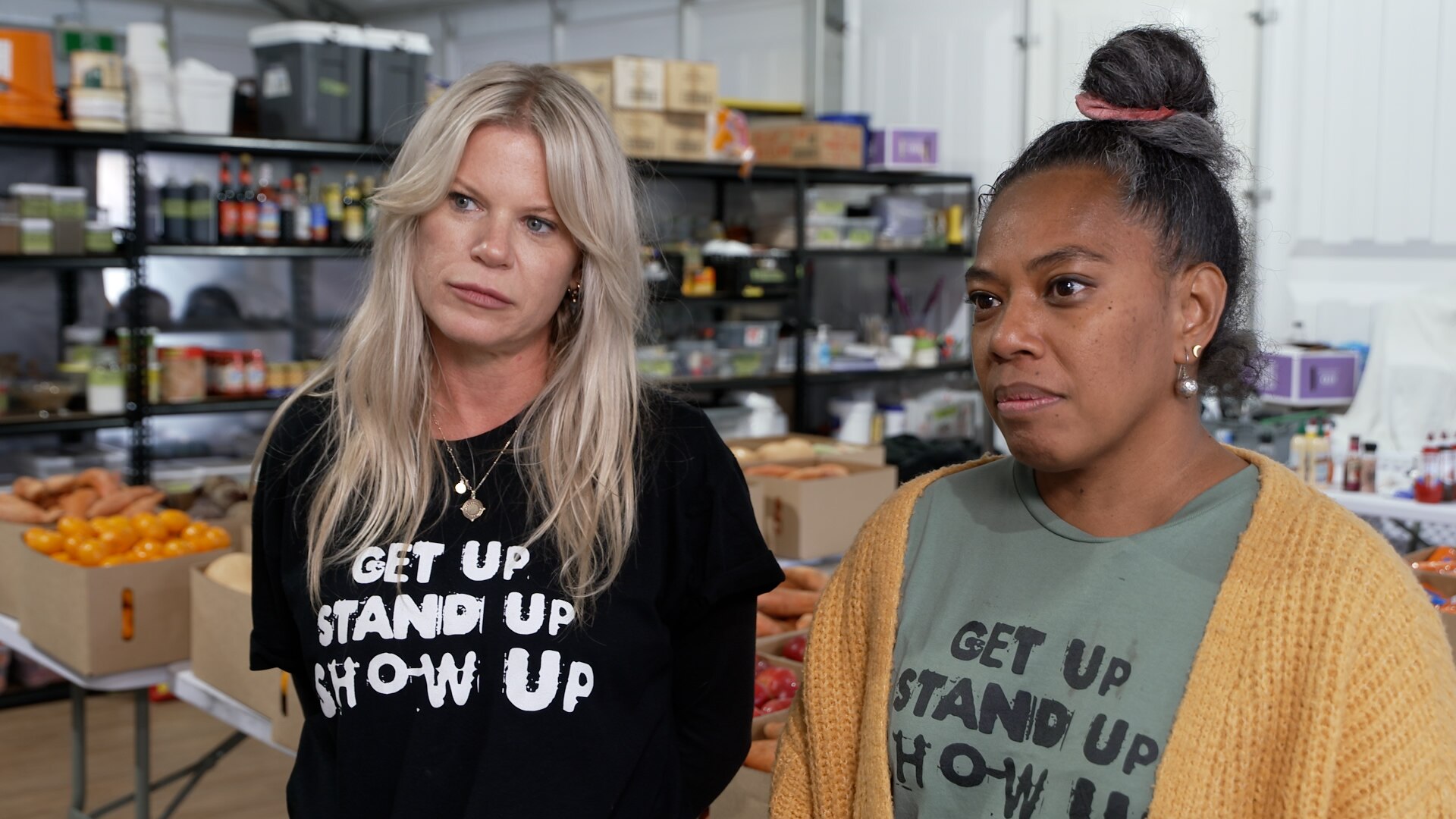 Two women standing together, one blonde one black hair, produce and shelves behind them.