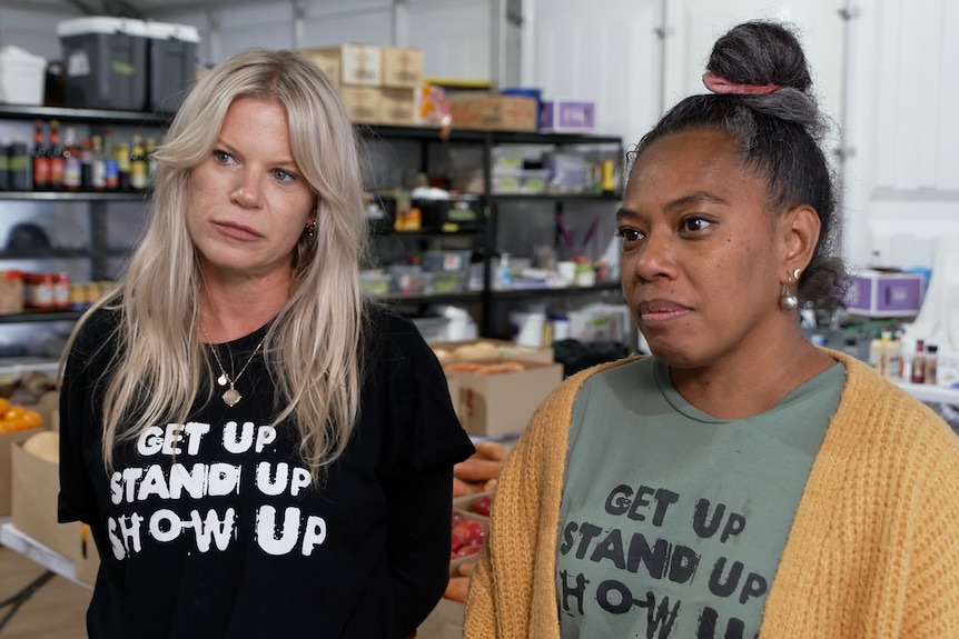 Two women standing together, one blonde one black hair, produce and shelves behind them.