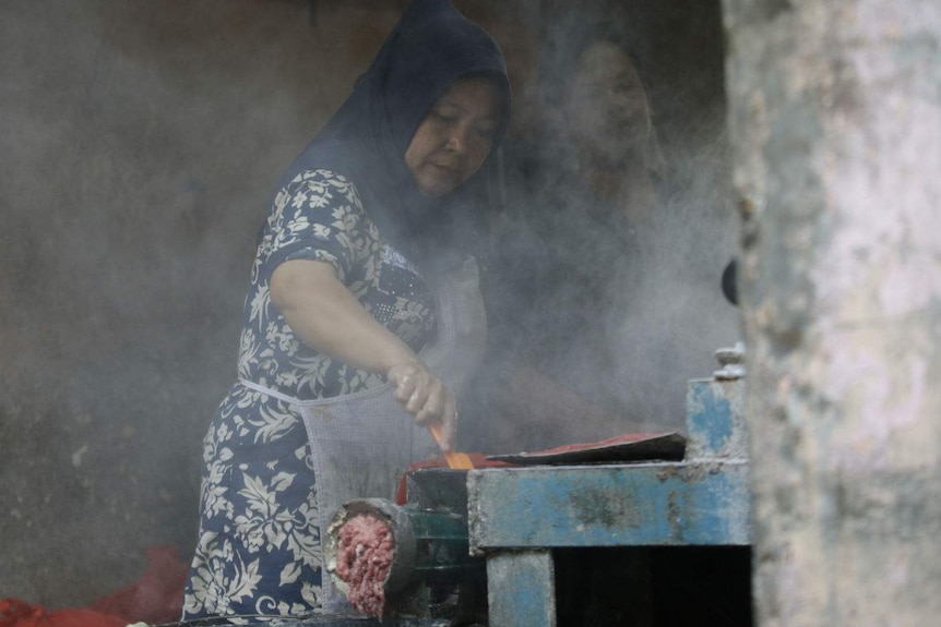 A woman in a hijab feeds meat through a mincer at a market stall.