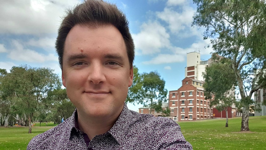 A man smiles at the camera with a grassy reserve and large factory in the background.