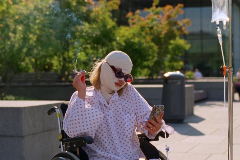 A woman with a bandaged face in a hospital gown, in a wheelchair, attached to a drip smokes a cigarette and checks her phone.