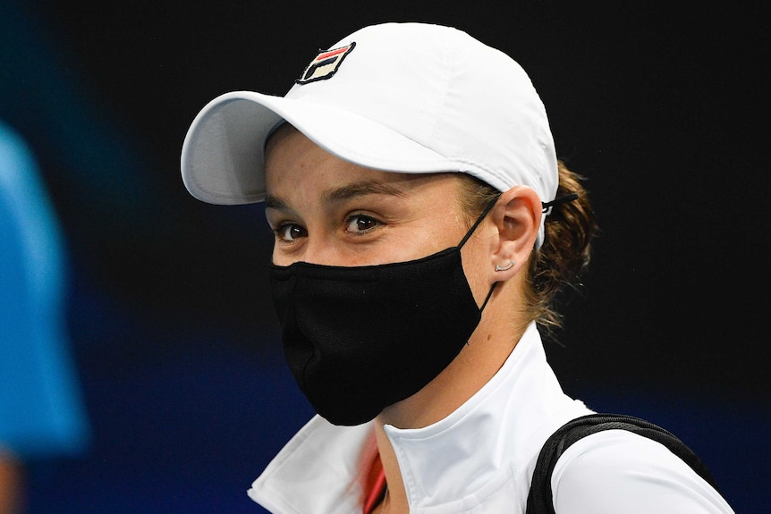A cap-wearing female tennis player reacts as she walks onto court wearing a mask.