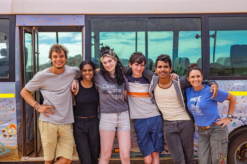 Six young people wearing tshirts and shorts stand together outside a colourful mini bus, smiling.