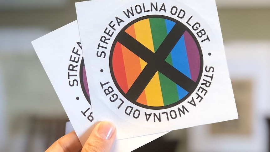 A hand is pictured holding two stickers with a black cross over a rainbow flag.