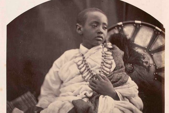 A young boy sits for a portrait in Britain alongside various items from Abyssinia. 