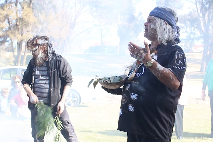 Two aboriginal men with body paint on and wearing scarves around their heads and holding eucalyptus leaves, surrounded by smoke