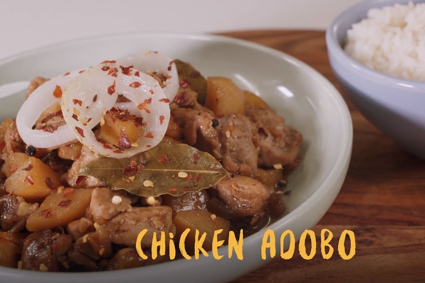 A white dish filled with chicken pieces, onion rings and potatoes. It's accompanied by a blue bowl of rice.