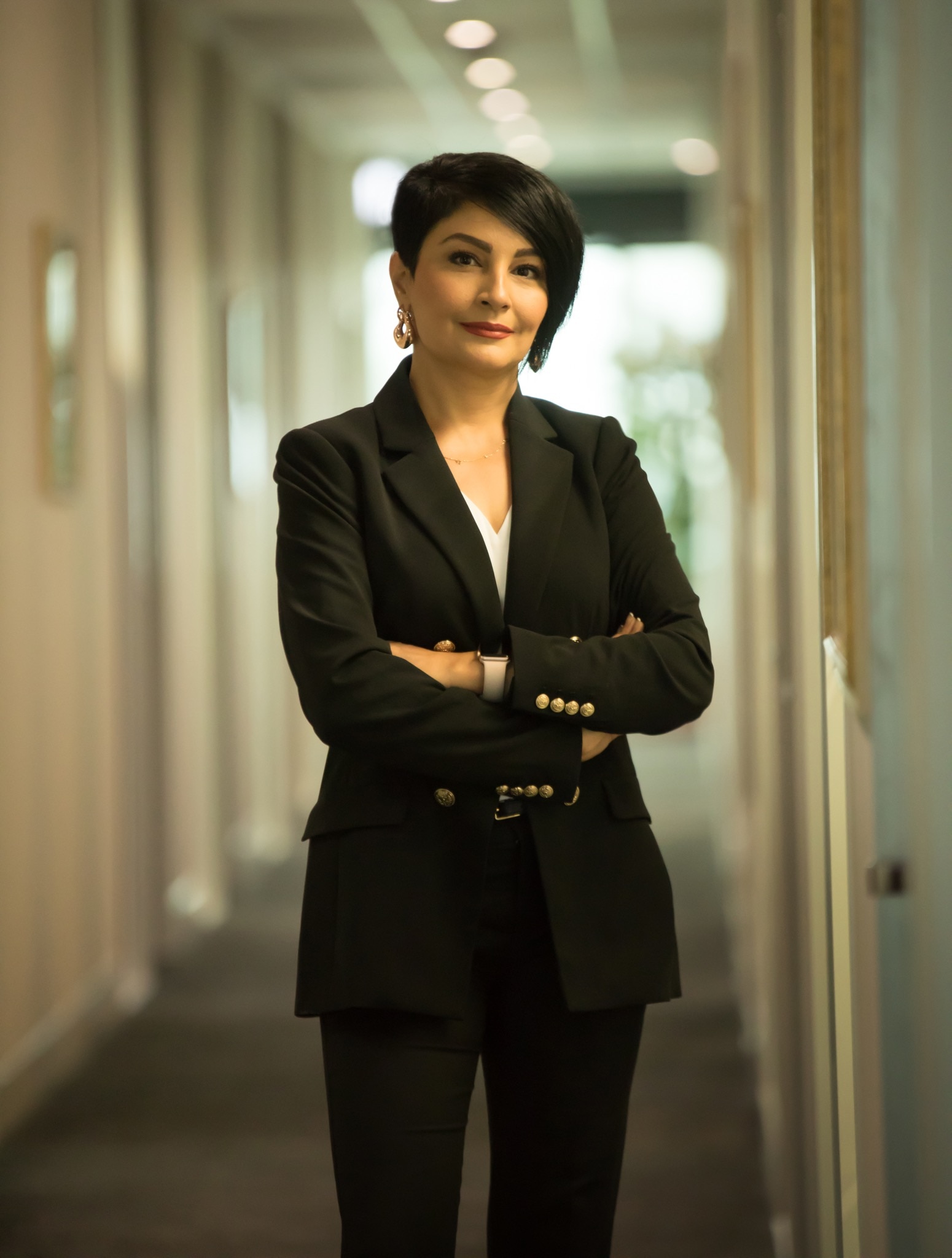 A well-groomed brunette woman, dressed for the office, smiles at the camera, her arms slightly crossed. 