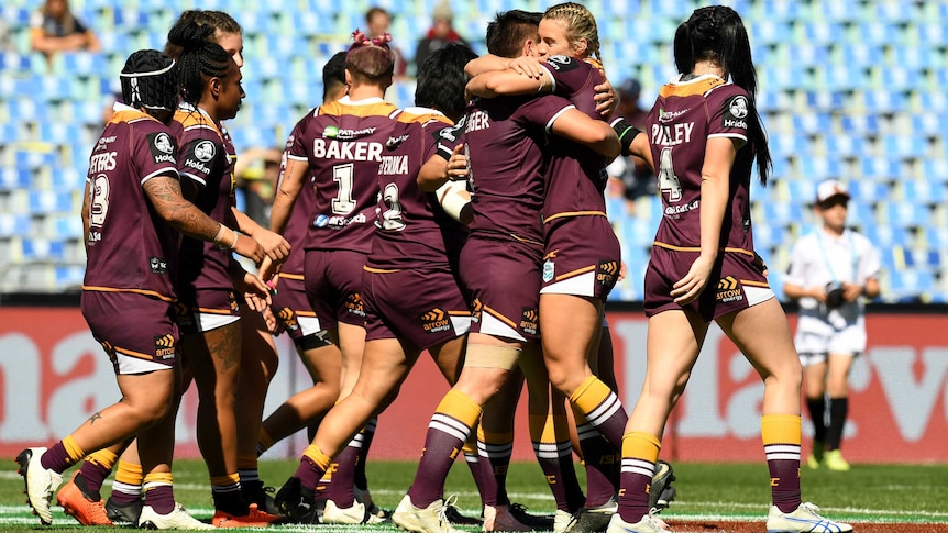 Women wearing marron jerseys hug in front of empty grandstands.