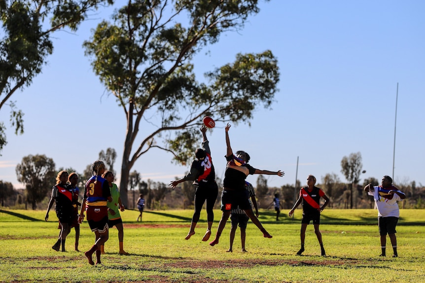 Two young Aboriginal women wearing football jumpers prepare to jump at a football thrown by an umpire
