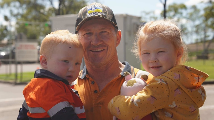 A man in a cap and high-vis smiles while holding two young kids.