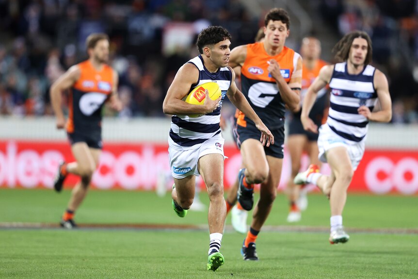 A Geelong AFL player holds the ball under one arm and looks to his left as he sprints away from a GWS player.