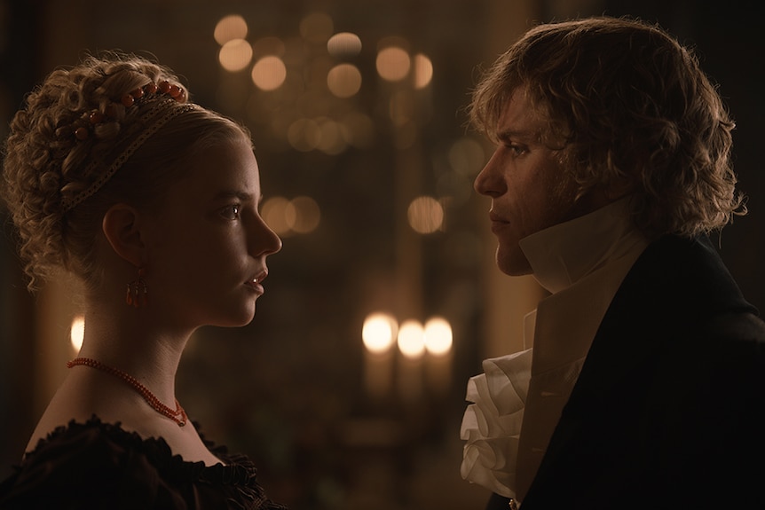 A blonde woman and man, both with curly hair, wear Regency England period costumes and stand intensely looking at each other.