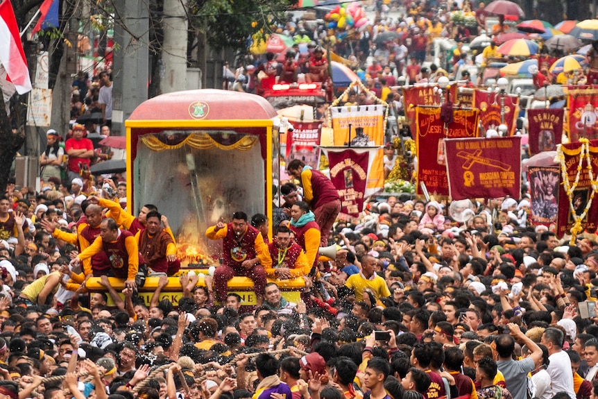 A crowd of devotees dressed in maroon and yellow climb a glass carriage  carrying the statue Black Nazarene.