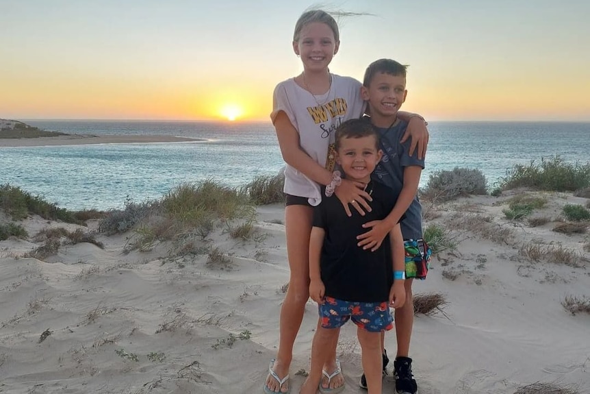 Three children stand on the beach with the sun setting behind them.