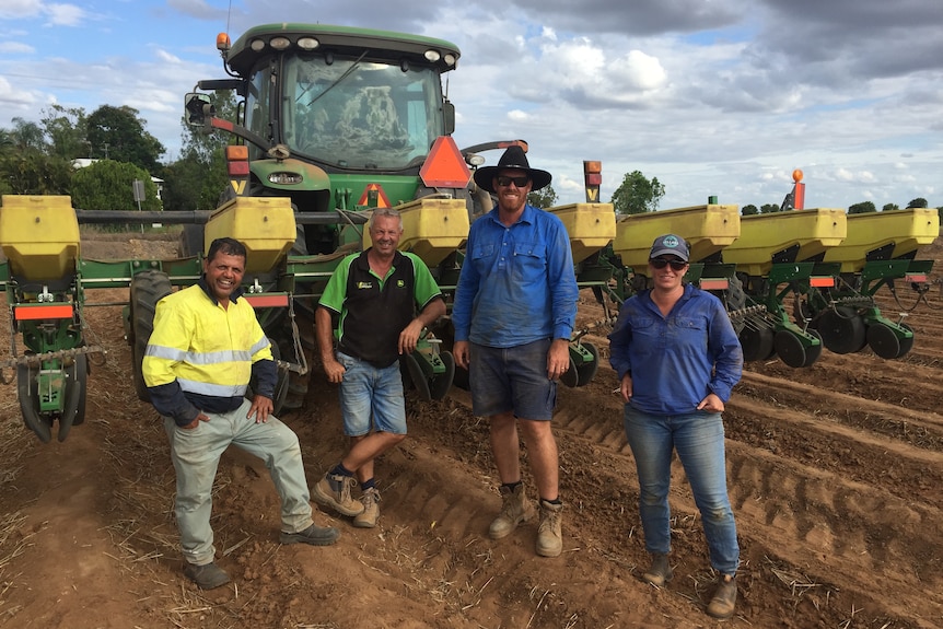 Four people stand next to eachother grinning in freshly dug rows in front of a large piece of machinery used for planting