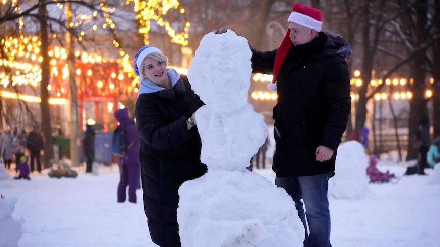 A man and woman build a snowman in a park covered with fairy lights. 