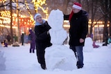 A man and woman build a snowman in a park covered with fairy lights. 