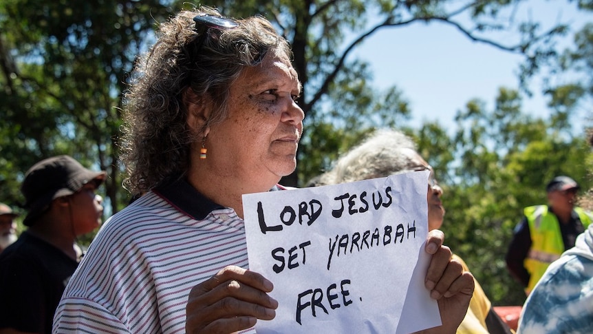 Indigenous woman holding sign that reads 'Lord Jesus set Yarrabah free'.