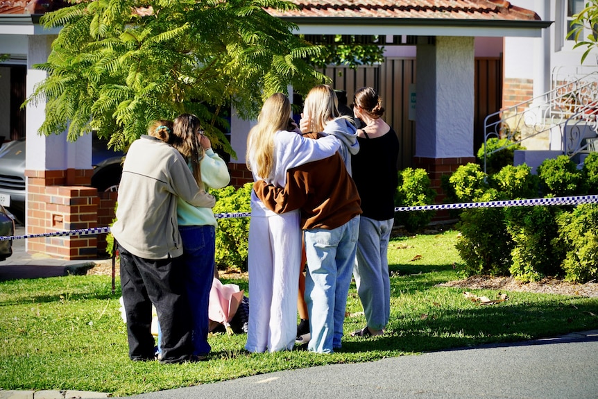 A group of five women stand huggled together outside a house.