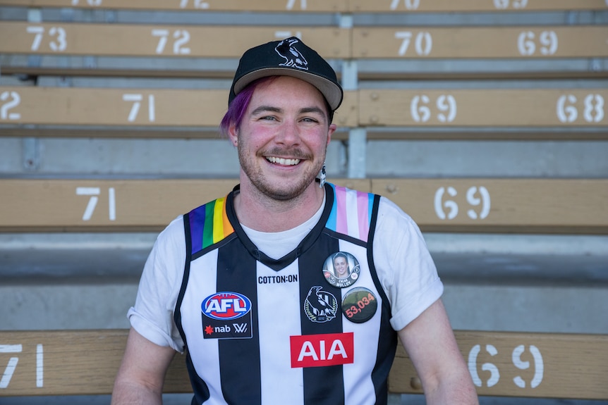Andy Pullar sits in a grandstand and smiles with a collingwood jersey on.